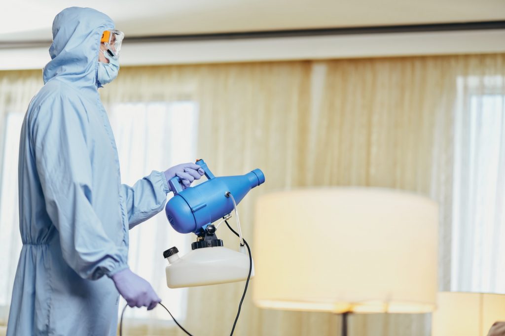 Man in blue protective suit holding disinfectant and standing in the room